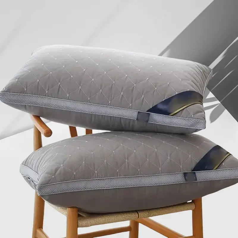 Pila de dos almohadas ortopédicas de estilo hotelero en color gris con diseño acolchado de rombos y banda lateral, sobre una silla de madera