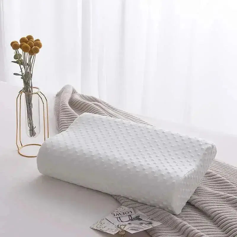 Vista de una almohada cervical blanca de memory foam con diseño de contorno ergonómico y textura de micro-puntos, presentada de forma elegante sobre una cama con una manta gris y un jarrón decorativo al fondo.